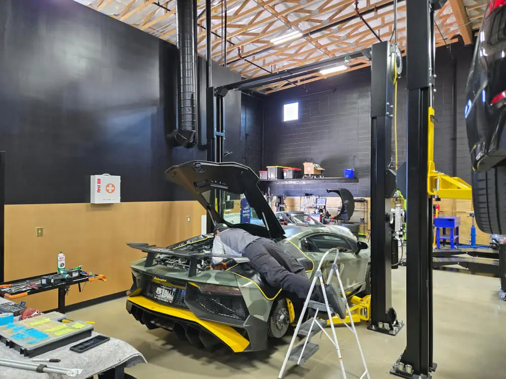 Aces Auto Elite - A mechanic lies on a platform, working under the open hood of a green sports car inside a well-lit garage. Tools and equipment are scattered nearby, and another car is elevated on a lift in the foreground. | Arizona Luxury & Exotic Car Service